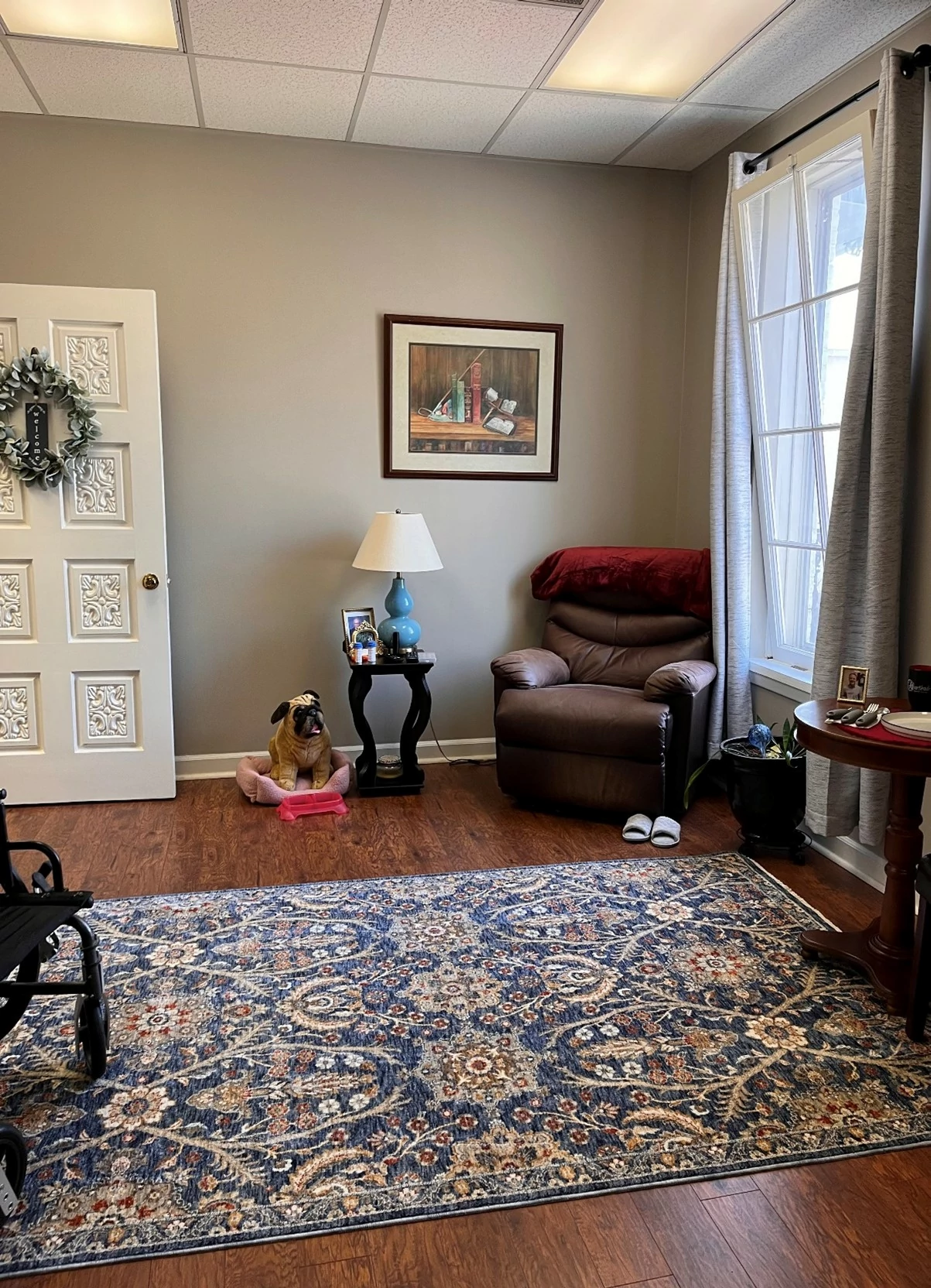 As you enter the studio apartment, you will be in the living space complete with a lift chair, Buddy the dog, and various fall risks.  To the right of the window, is a small kitchen table set with weighted utensils and other items to aid in training.